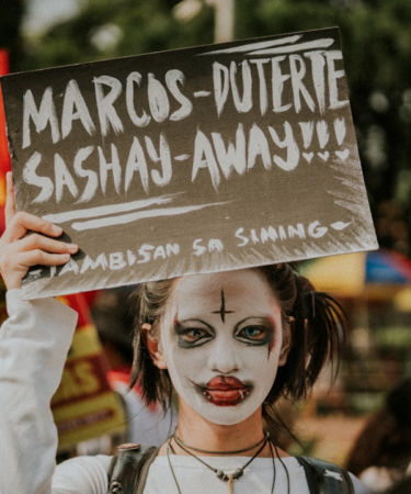 Person with clown makeup holds protest sign — Foto por Joaquin Arenas na Unsplash