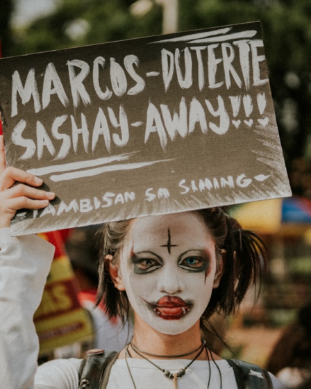Person with clown makeup holds protest sign — Foto por Joaquin Arenas na Unsplash